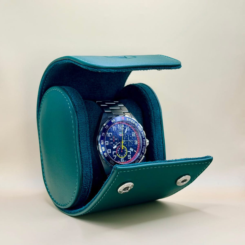 A teal leather watch display case with a metal snap closure, shown open with a wristwatch inside on a light beige background.