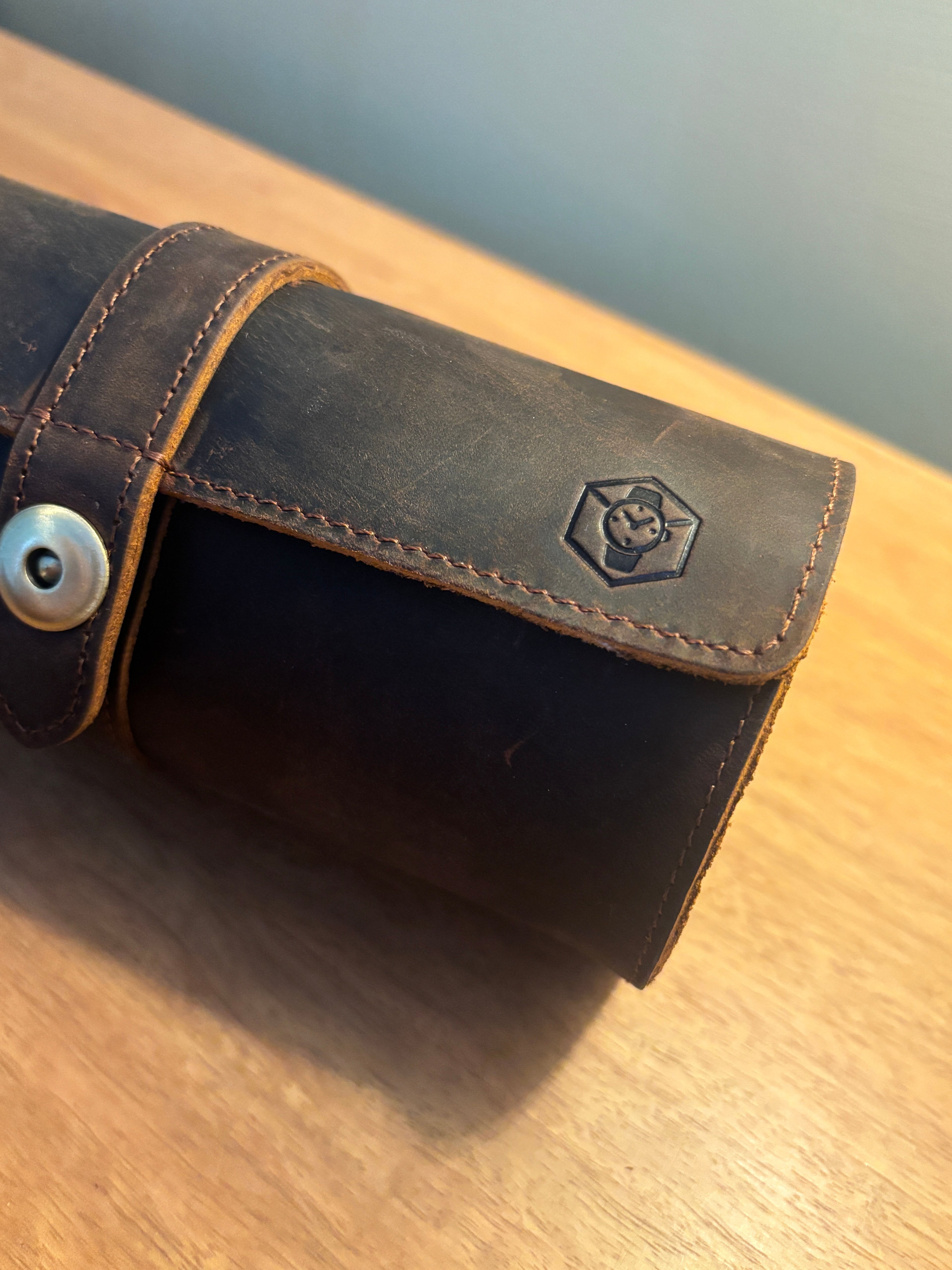 A close-up of a dark brown leather accessory, possibly a wallet or sleeve, featuring a snap-button closure and an embossed logo on the flap, resting on a wooden surface.