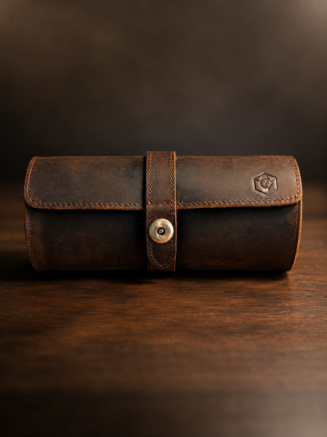 A dark brown distressed leather cylindrical case with a strap closure and metal snap button, resting on a wooden surface against a dark brown background.